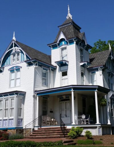 Front view of Laurel Manor House Bed & Breakfast in Laurel, Maryland — a restored 1888 Victorian home with ornate architectural detailing, central turret, blue and lavender trim, and a wraparound porch, framed by gardens and trees under a clear blue sky.
